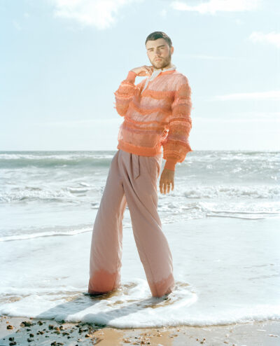 Male model dressed in androgynous clothing. He is stood in the sea, surrounded by water, while facing the camera.
