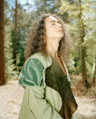 Male model stood in the middle of the forest, bathed in sunlight. He is looking up, while his eyes are shut.