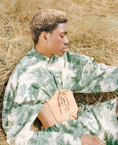 Male fashion model dressed in a timeless graphic tunic and a corset is laying down in a field. He closes his eyes in tranquility.