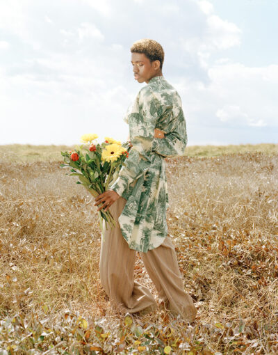Male model stood in the middle of a dry, sunbathed field. He looks of camera, while holding a bouquet of flowers.