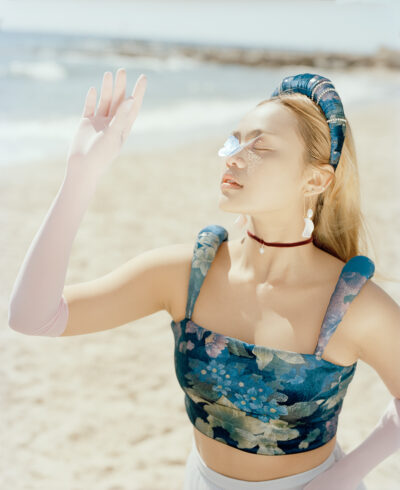 This is a photograph taken as a part of a fashion series. It features a female model stood at the beach with her eyes closed and hand raised. There is a metal butterfly on her nose.
