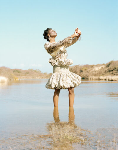 A fashion photograph showcasing a female model stood in a pond, while looking up with eyes closed. Her body is reflected in the water.