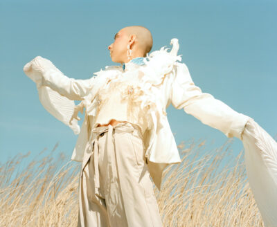 Model looks away from the camera, while raising both her arms resulting in the jacket sleeves blowing in the wind. This fashion editorial image is taken amongst plants, against a clear blue sky.