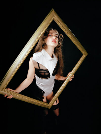 Female model stood in the photo studio, against a black backdrop holding a golden picture frame around her looking at the camera