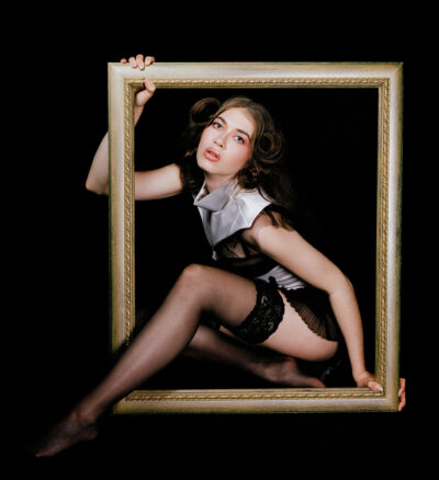 An old painting style photograph of a female actress dressed in Victorian lingerie looking up at the camera as she pokes her leg through a golden picture frame