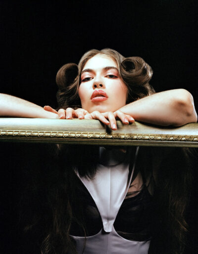 Portrait of a model dressed in modern Victorian fashion stood against a black studio backdrop as she leans onto a golden picture frame