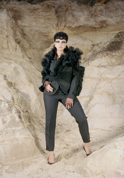 Female model stood tall against a cliff background looking down at the camera powerfully, wearing a black custom suit and headpiece with golden elements