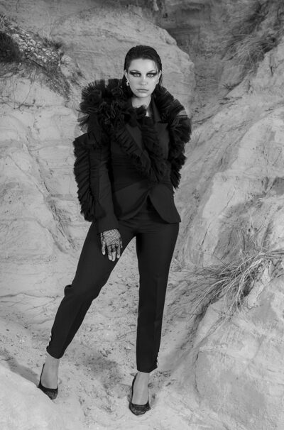 monochrome image of a female model dressed in a designer suit with ruffles, stood fiercely in front of a cliff, looking at the camera