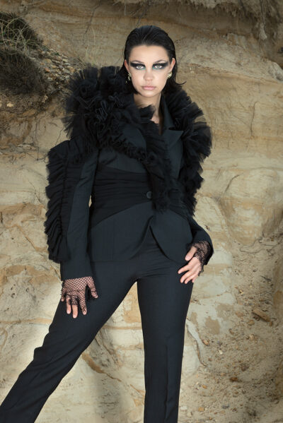 Female model stood against a cliff holding one of her hips in one hand, wearing a custom made black suit for a fashion shoot